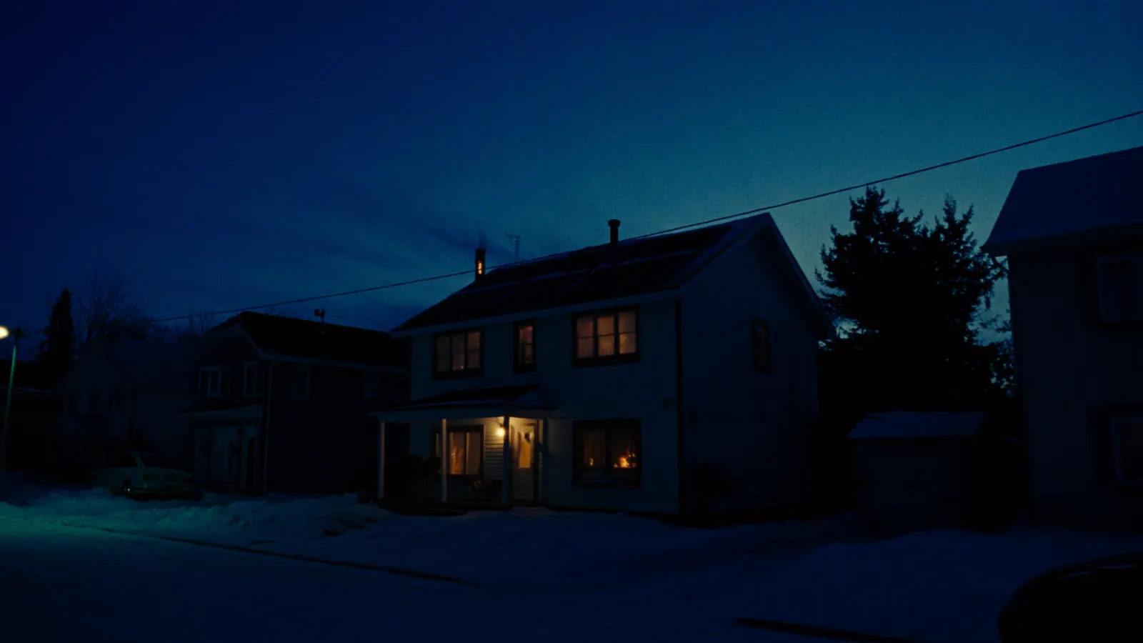 A snowy suburban street at night during an outage, every house dark except one with warm lit windows and solar panels on its roof